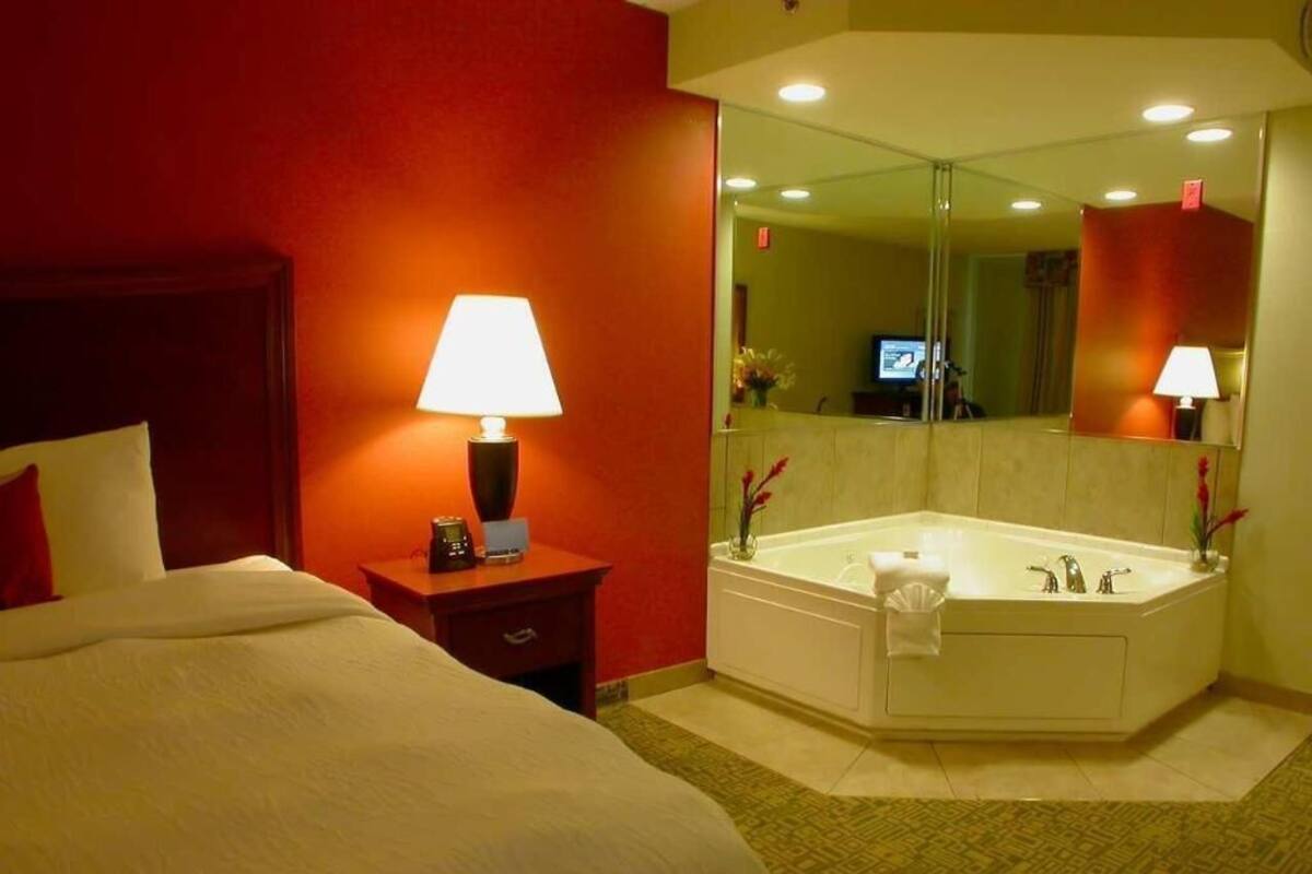 A hotel room with a bed, a nightstand with a lamp, a large mirror, and a bathtub.