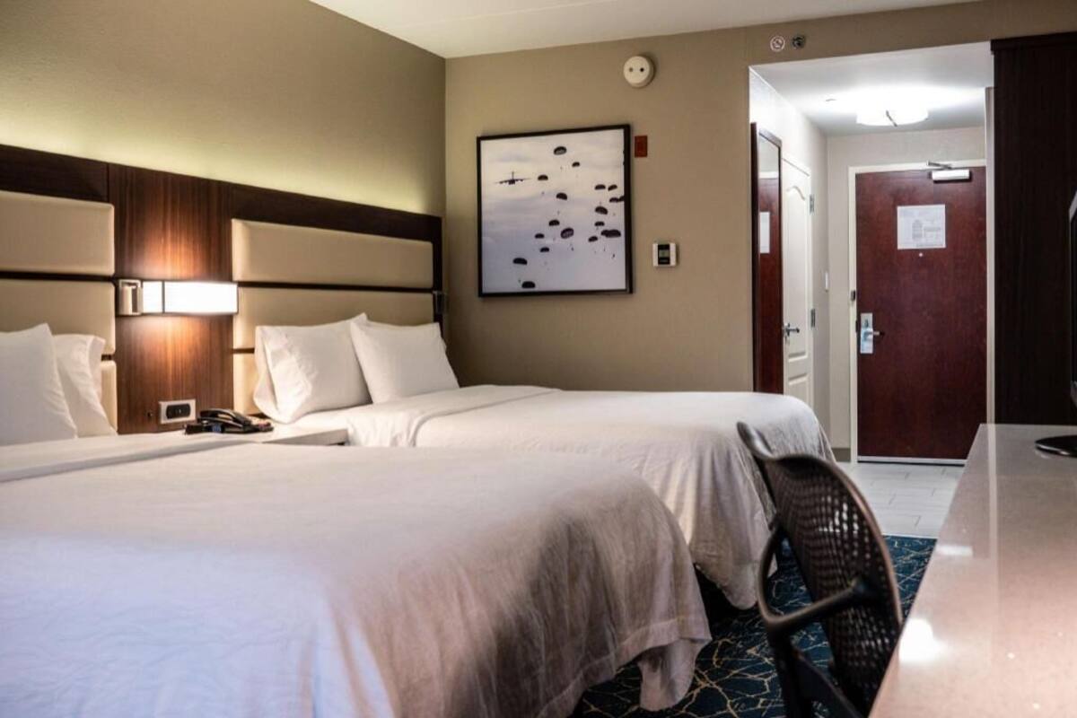 A hotel room with two beds, a headboard with integrated lighting, a wall-mounted artwork, a desk with a chair, and a door leading to another room.