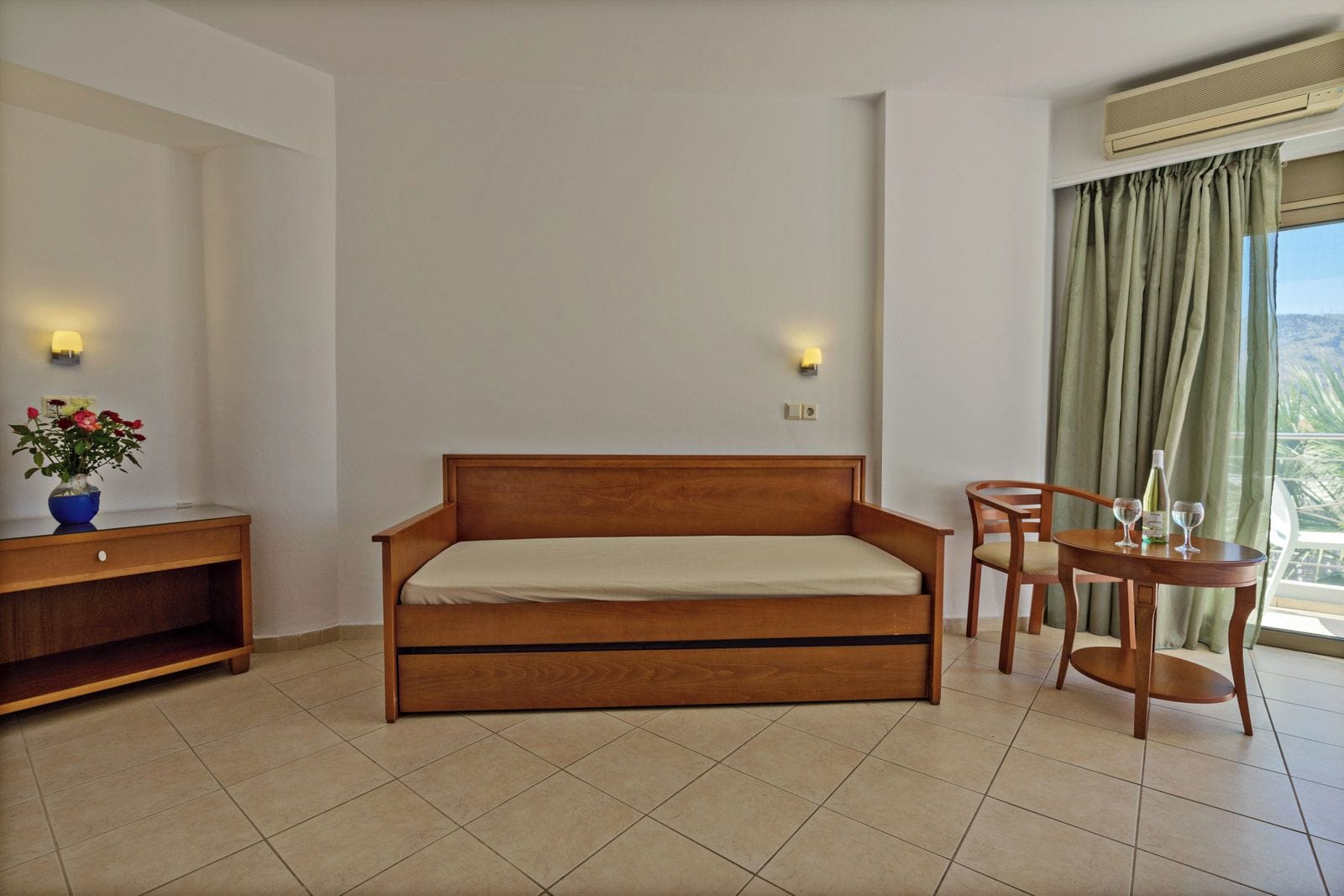 A room with a wooden bed, a small table with two chairs, a vase with flowers, and a window with curtains.