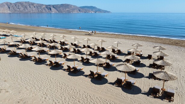 A beach with rows of sun loungers and thatched umbrellas, a clear sky, and distant mountains. - Delfina Boutique (Apokoronas)