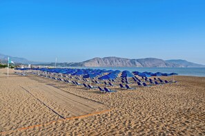 A beach with rows of blue umbrellas and sun loungers, a flag, and mountains in the background. - Delfina Boutique (Apokoronas)