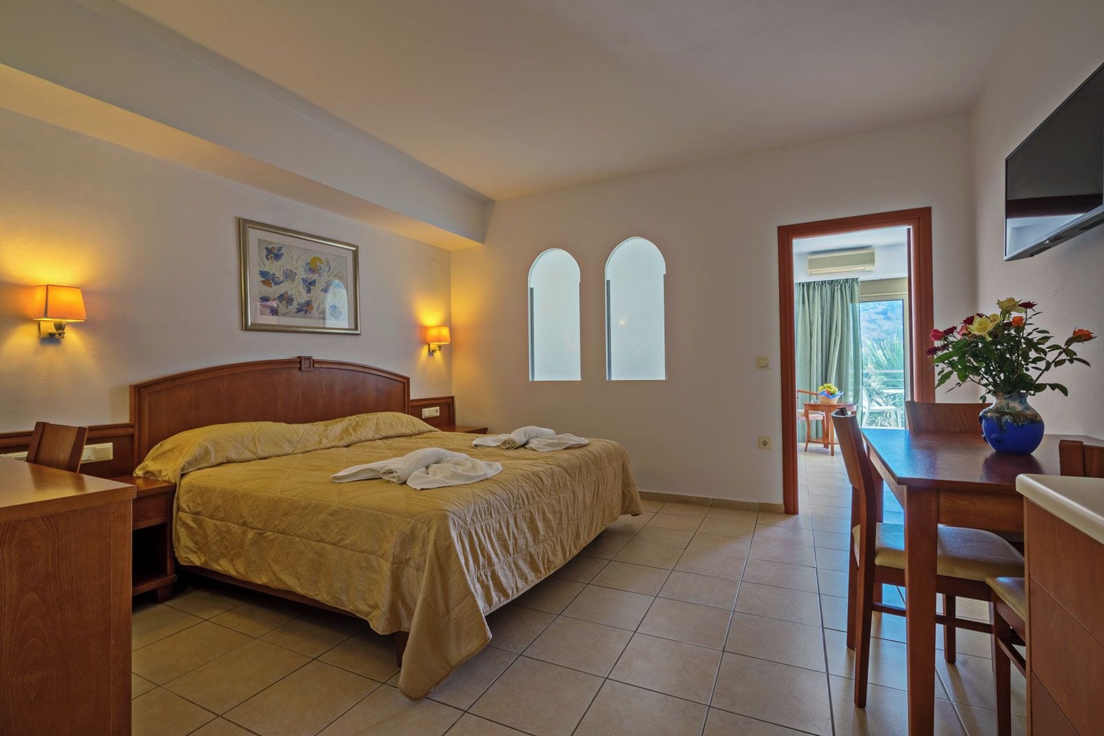Una habitación de hotel con cama, mesitas de noche, escritorio, silla, televisor, un cuadro y ventana.