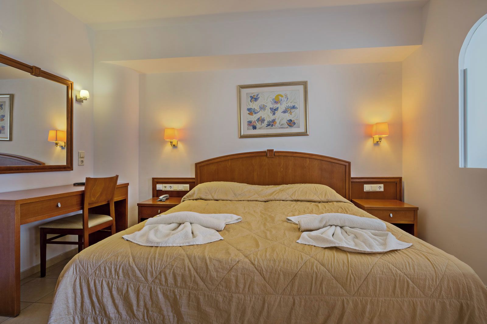 Habitación de hotel con cama, dos mesitas de noche, escritorio con silla, espejo y dos lámparas de pared.