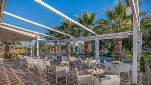 An outdoor cafe with white wicker furniture, a tiled floor, and palm trees in the background. - Delfina Boutique (Apokoronas)