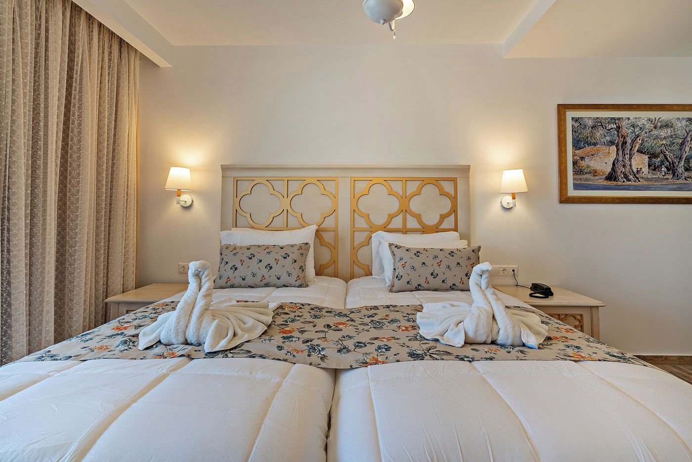 Une chambre d’hôtel avec un grand lit, deux tables de chevet, une tête de lit ornée d’un panneau décoratif, des lampes fixées au mur et un tableau accroché au mur.