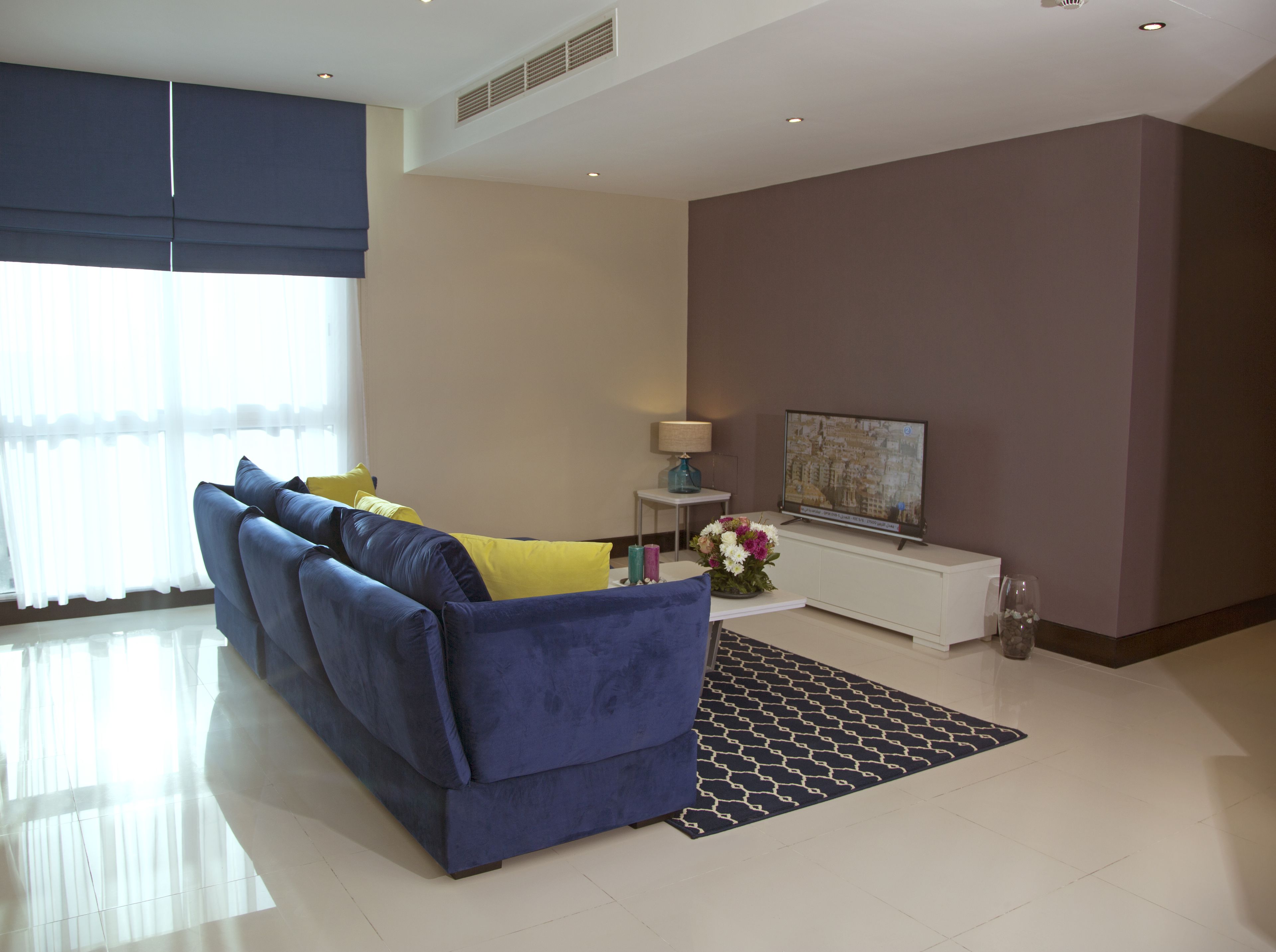 A modern living room with a blue sofa, a small side table with a lamp, a flat-screen TV, and a decorative vase.