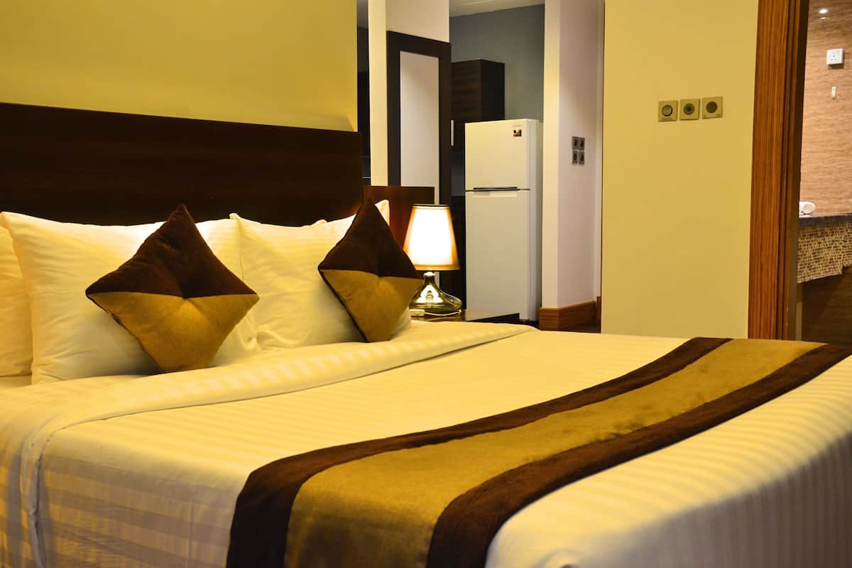 A hotel room with a large bed, a nightstand with a lamp, and a refrigerator in the background.