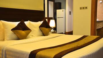 Una habitación de hotel con una cama grande, una mesita de noche con lámpara y un refrigerador al fondo.