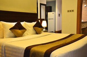 Una habitación de hotel con una cama grande, una mesita de noche con lámpara y un refrigerador al fondo.