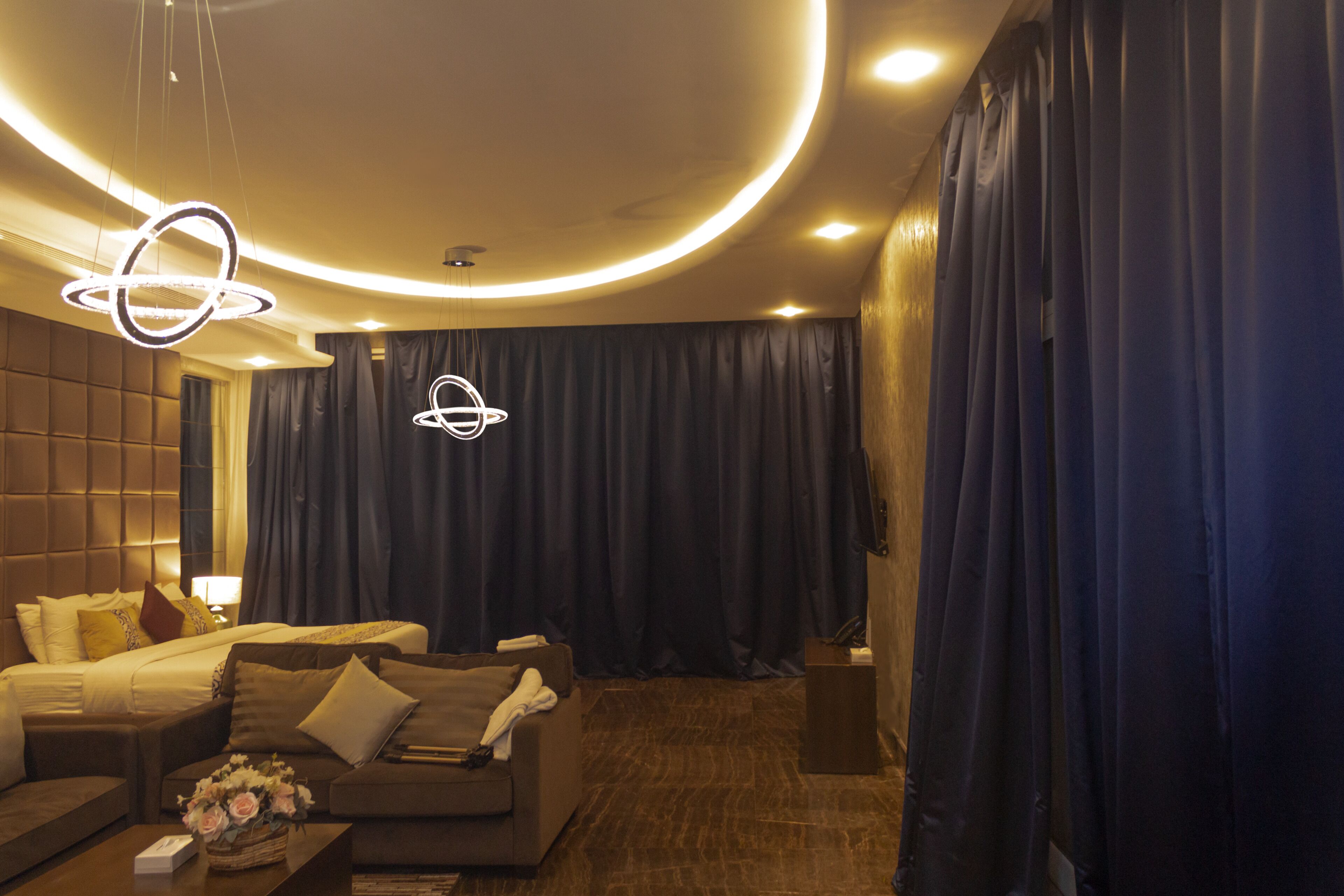 Una habitación de hotel moderna con una cama grande, un sofá y una mesa de centro. Hay cortinas, una araña y un televisor montado en la pared.