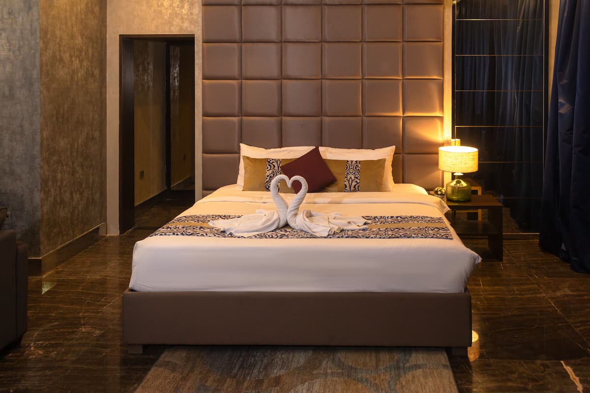 A hotel room with a large bed, a headboard with a padded design, a bedside table with a lamp, and a nightstand with a green object.
