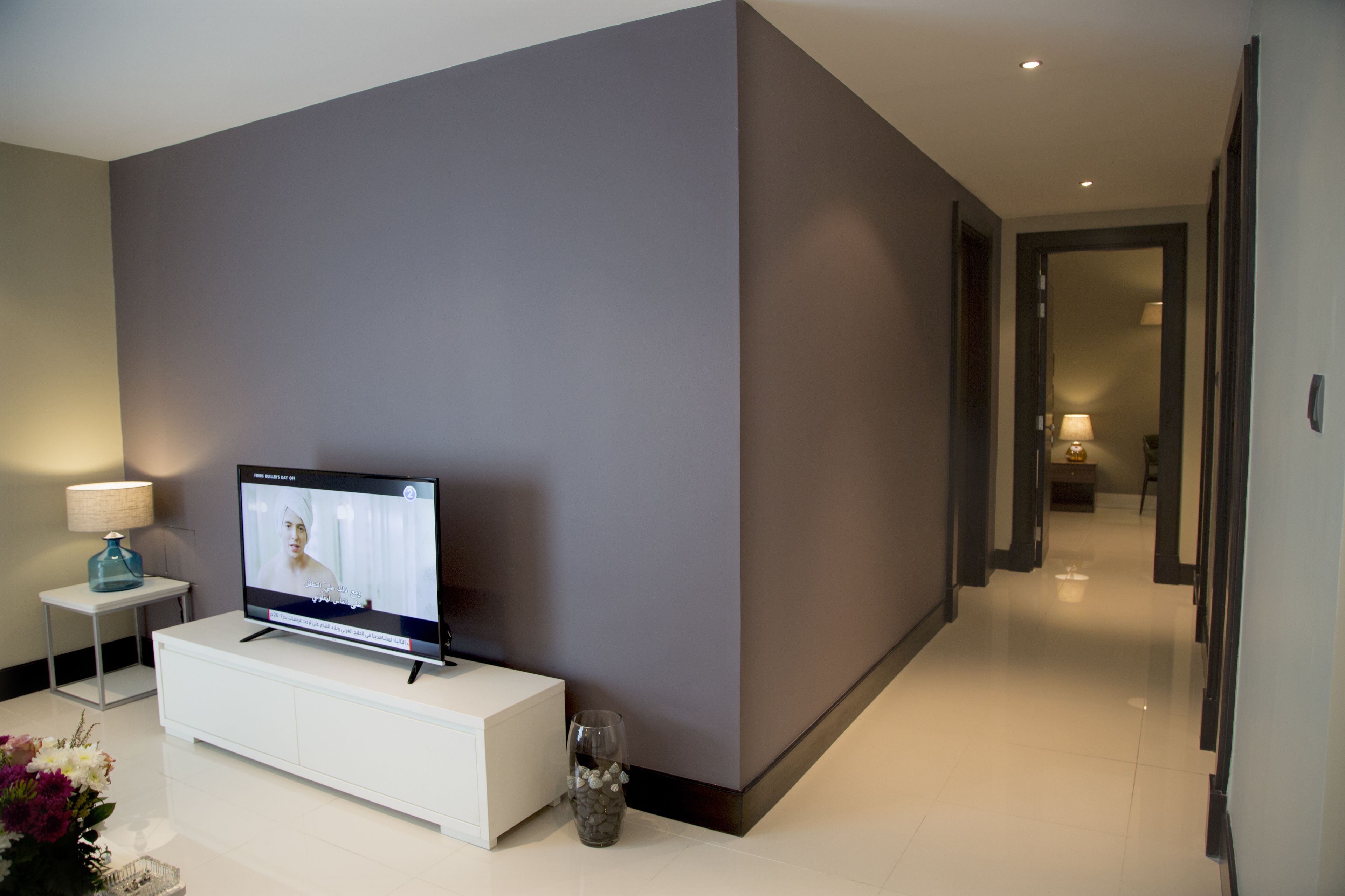 Habitación de hotel moderna con un televisor de pantalla plana sobre una consola blanca, una mesita con una lámpara y un florero con piedras.