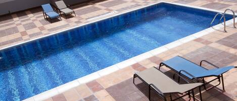 Una piscina rectangular con un borde de azulejos, dos tumbonas y una mesa de metal con sillas.