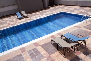 Una piscina rectangular con un borde de azulejos, dos tumbonas y una mesa de metal con sillas.