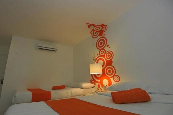 Una habitación de hotel con una cama, almohadas anaranjadas, una lámpara de pared y un equipo de aire acondicionado.
