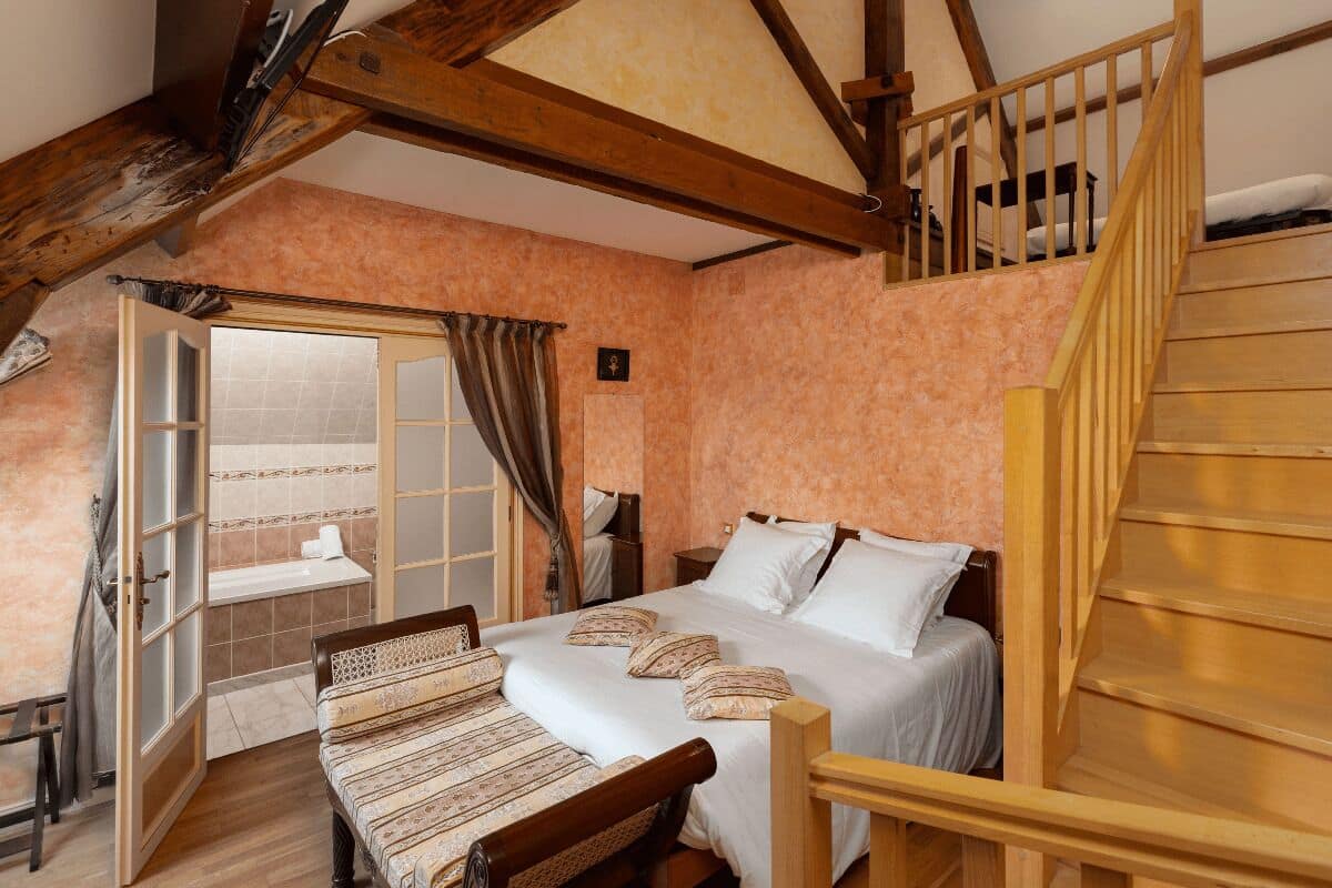 Um quarto com cama e escada de madeira, uma janela com cortinas e porta de acesso a um banheiro.