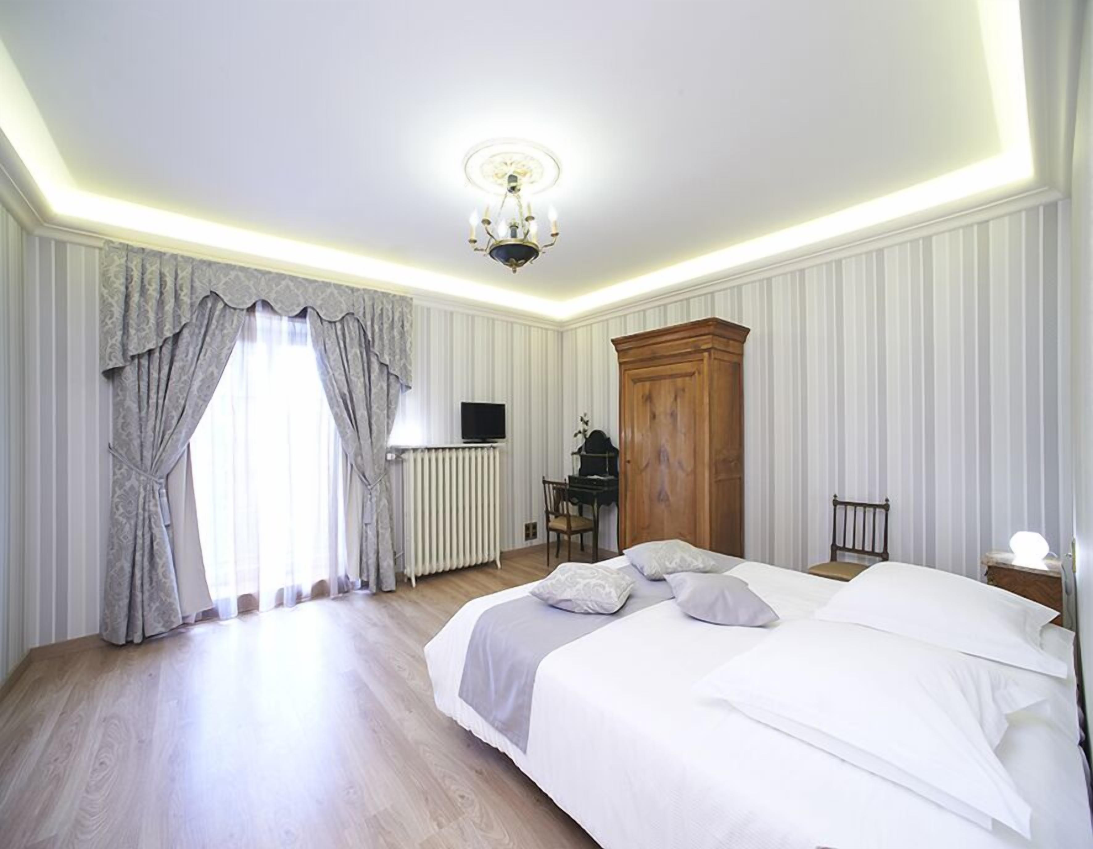 A bedroom with a large bed, a wooden wardrobe, a radiator, a television, and a chandelier.