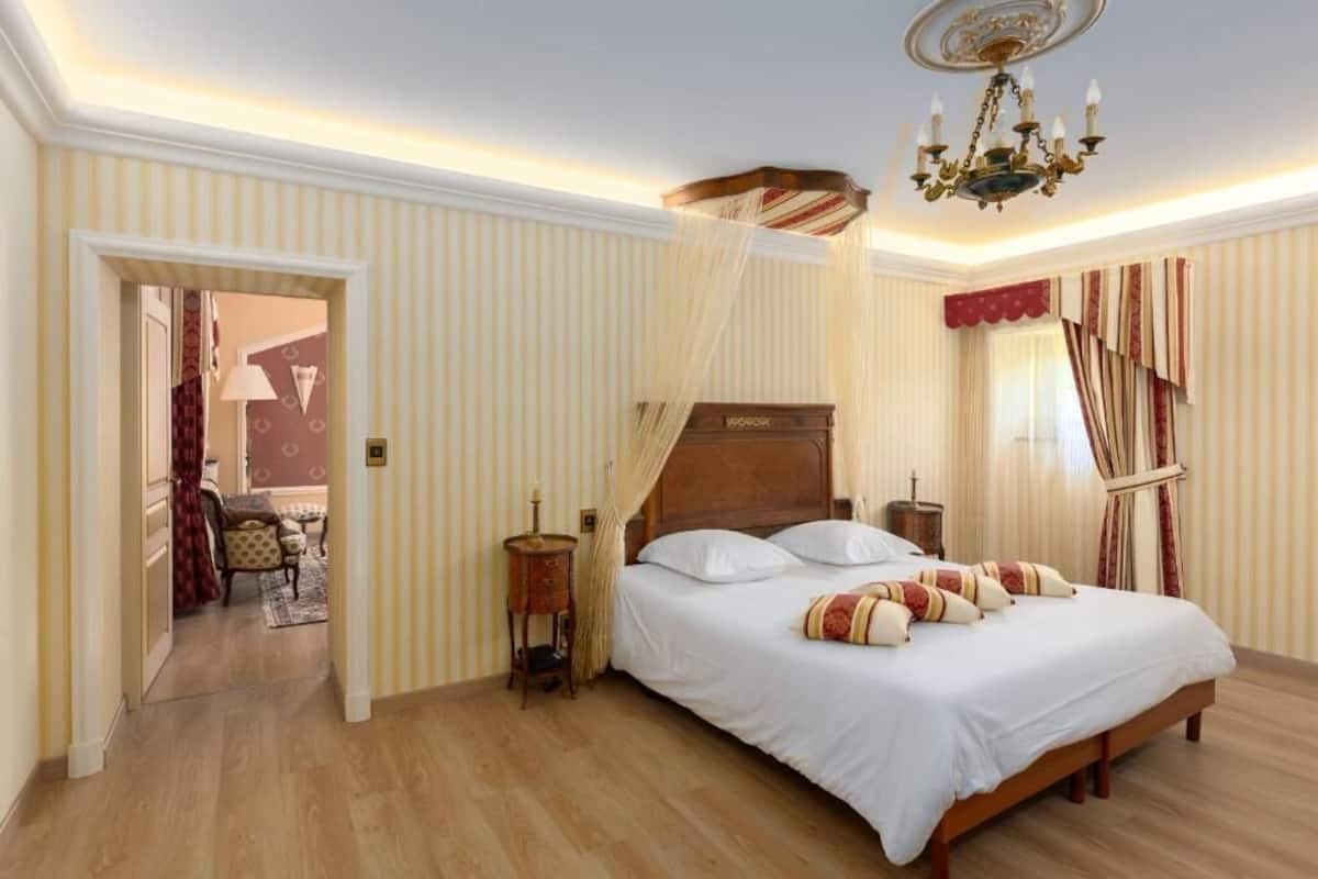 Quarto com cama grande, cabeceira de madeira, lustre e vista para uma sala de jantar através de uma porta aberta.