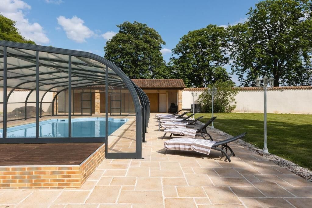 A swimming pool with a glass enclosure, lounge chairs, and a brick patio.