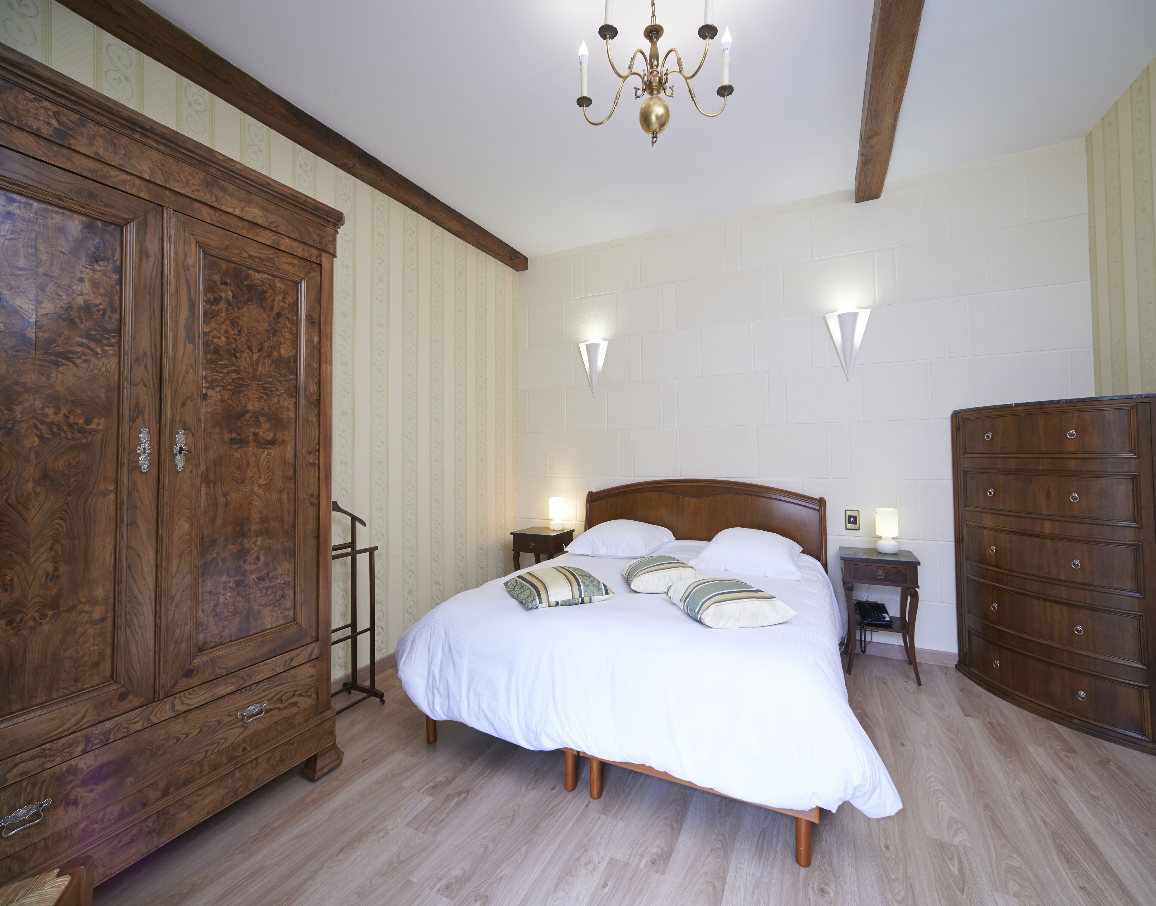 Une chambre avec une grande armoire en bois, un lit en bois avec une literie blanche, deux tables de chevet avec des lampes et un lustre.