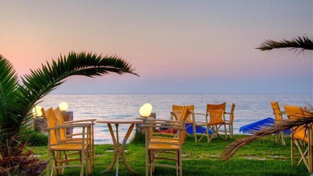 An outdoor seating area with wooden chairs and tables, overlooking a body of water. - Krini Beach Hotel (Rethymno)