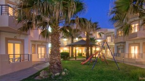 A hotel courtyard with palm trees, a playground, and a covered seating area. - Krini Beach Hotel (Rethymno)