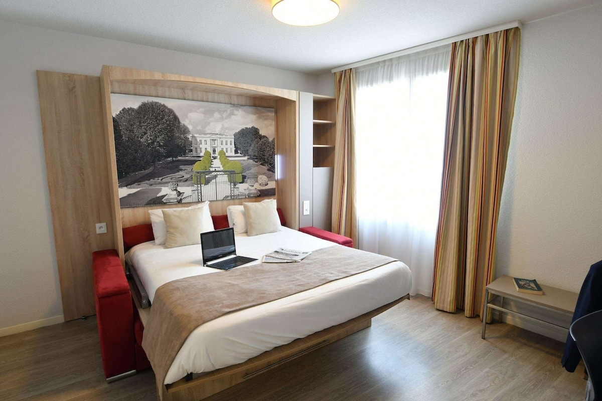 Ein Hotelzimmer mit einem Bett, einem Laptop, einer roten Bank, einem Bücherregal und einem großen Fenster mit Vorhängen.
