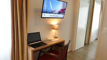 Ein Hotelzimmer mit Schreibtisch, Laptop, Stuhl, Fernseher, Lampe und Vorhängen.