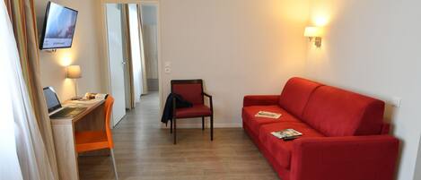 A hotel room with a wooden desk, a red sofa, a TV mounted on the wall, and a lamp.
