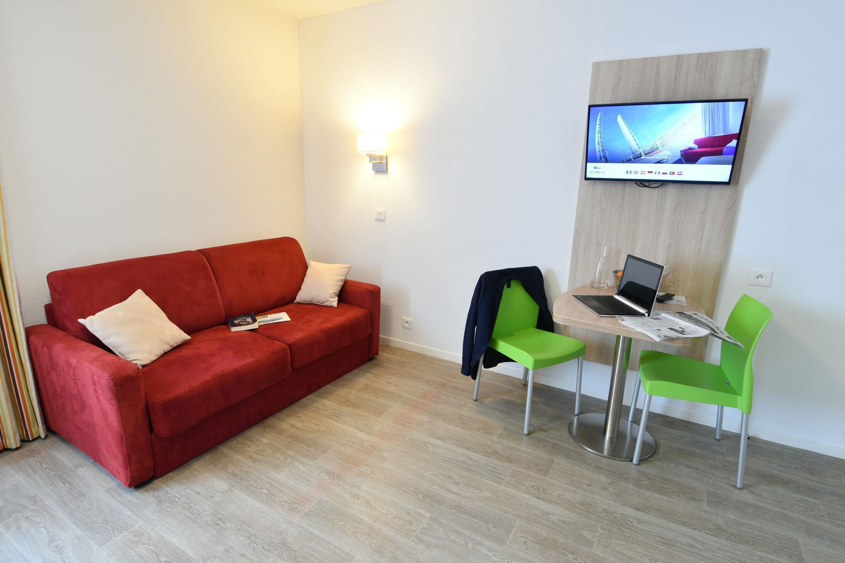 Una habitación con un sofá rojo, dos sillas verdes, un escritorio de madera con una computadora portátil y un televisor montado en la pared.