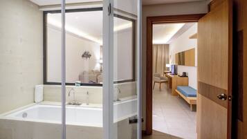 A modern bathroom with a large bathtub, a glass shower enclosure, and a wooden door leading to a bedroom with a bed and a desk.