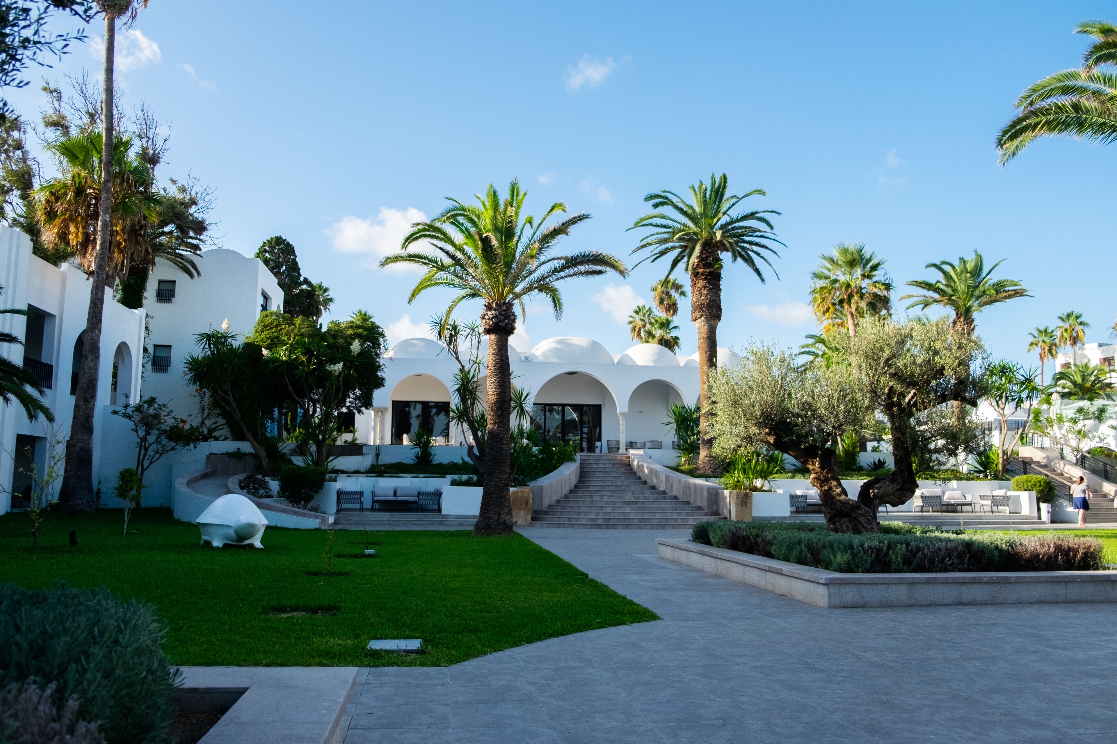 Un patio con palmeras, un edificio blanco con arcos y una escalera que conduce a la entrada.
