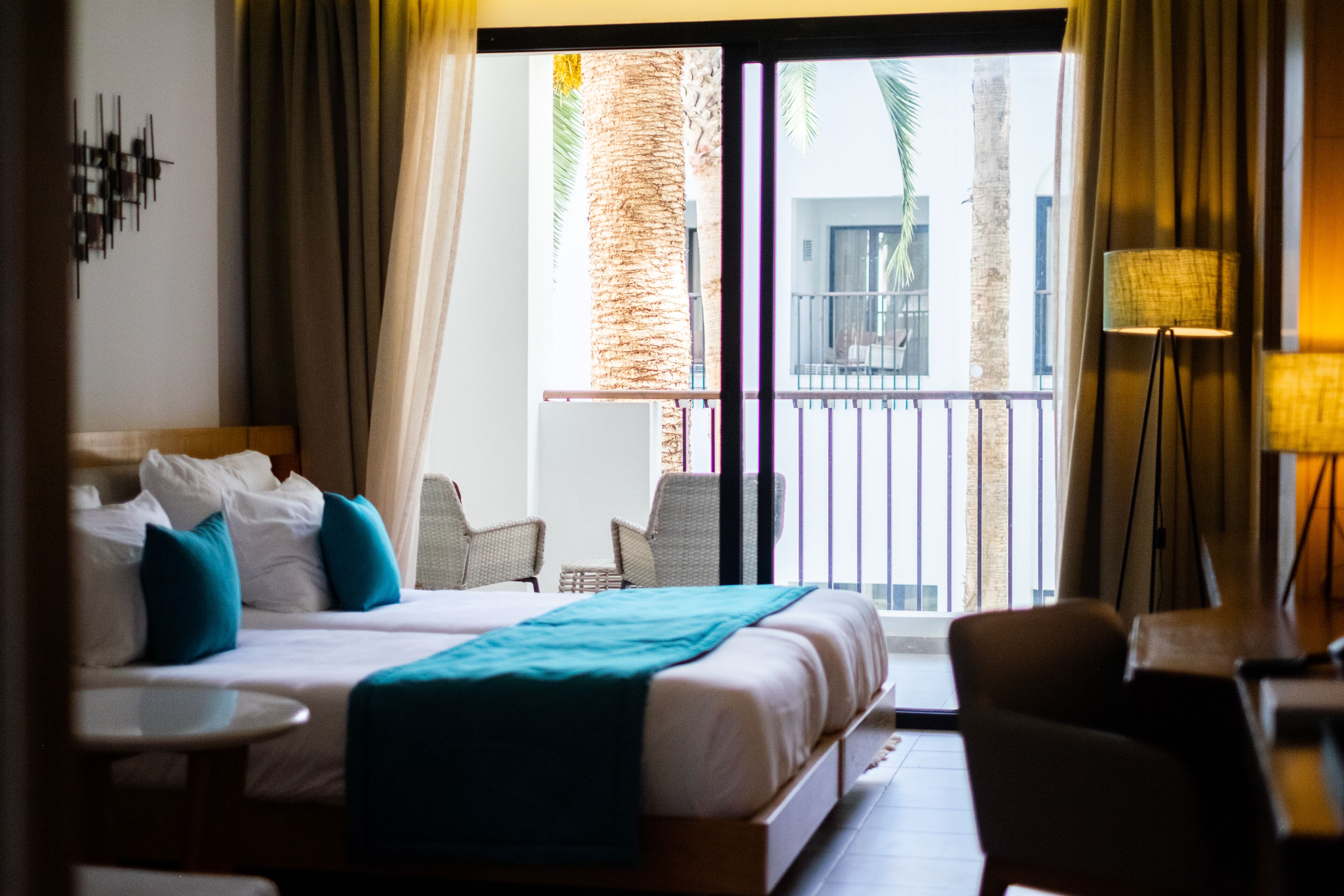Habitación de hotel con cama, una mesita, una silla y balcón con vistas a palmeras y edificios.