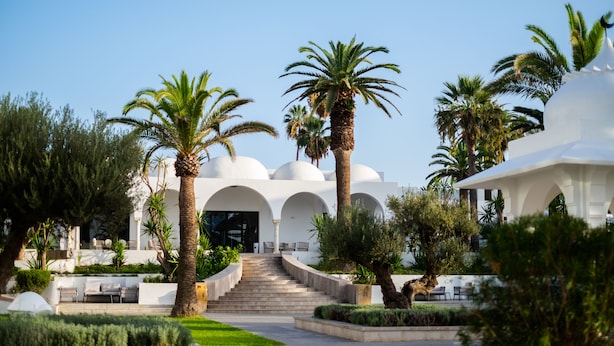 Een resort met een centrale trap die leidt naar een wit gebouw met bogen, omringd door palmbomen en groen.