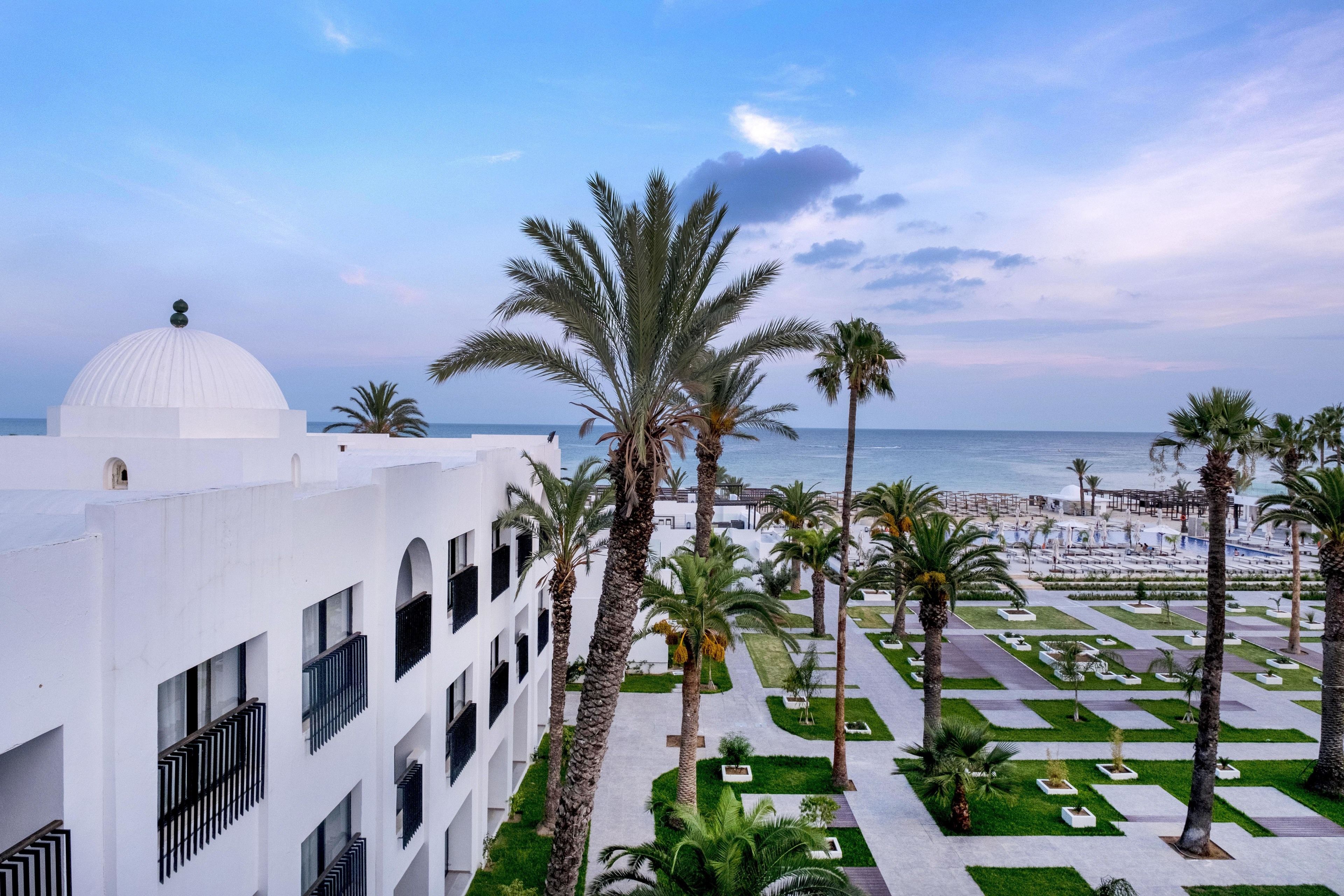 A hotel with a white facade, palm trees, and a view of the beach.