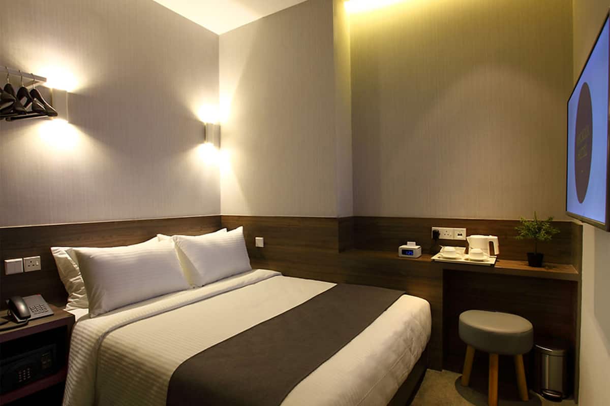 A hotel room with a neatly made bed, bedside table with a lamp, a small stool, and a wall-mounted TV.