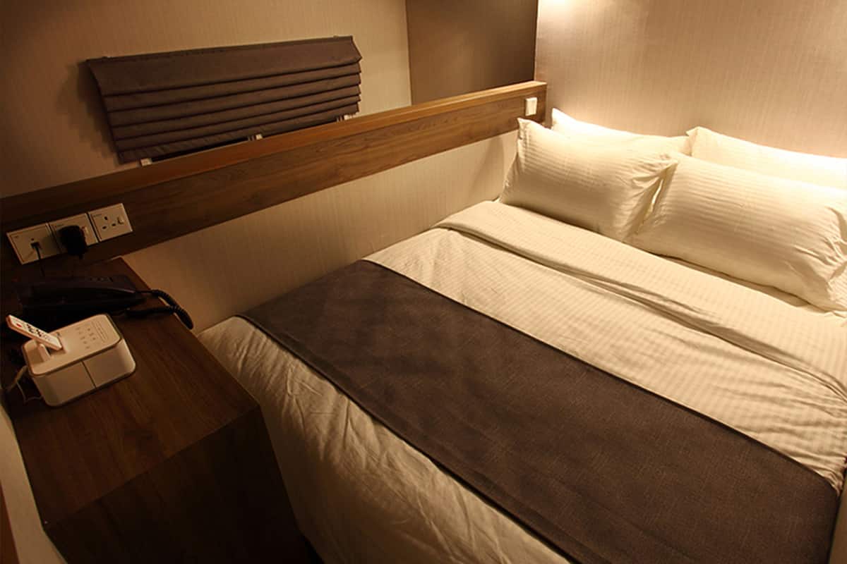 A hotel room with a bed, a bedside table with a phone, and a wall-mounted shelf.