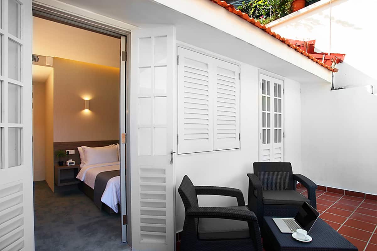 A hotel room with a bed, a seating area with two chairs, a small table with a laptop and a cup, and a balcony with a red railing.