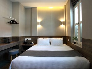 A hotel room with a large bed, bedside tables, a desk, a chair, and a window with blinds.