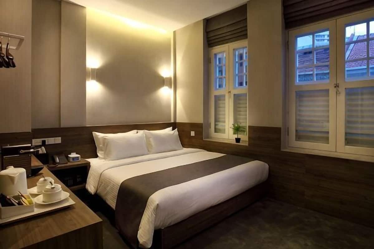 A hotel room with a large bed, bedside tables, a window with blinds, and wall-mounted lighting.