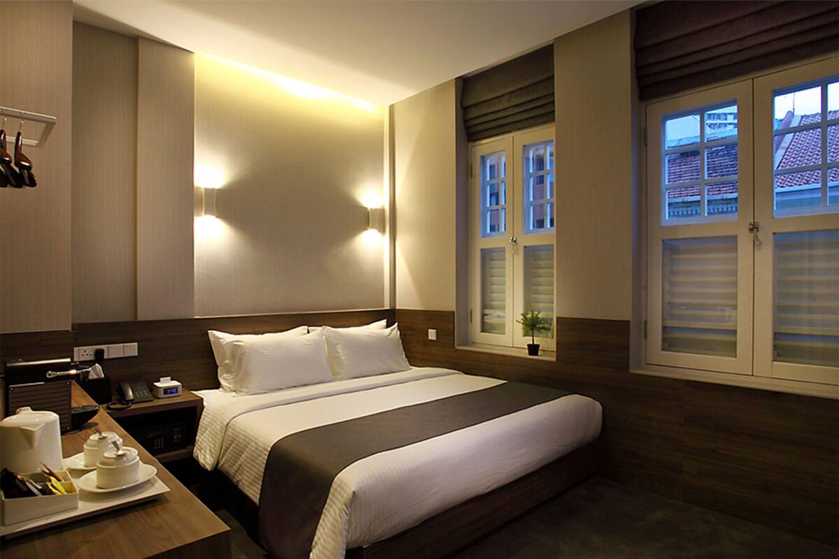 A hotel room with a large bed, bedside tables, a desk with a chair, a small potted plant, and a window with blinds.