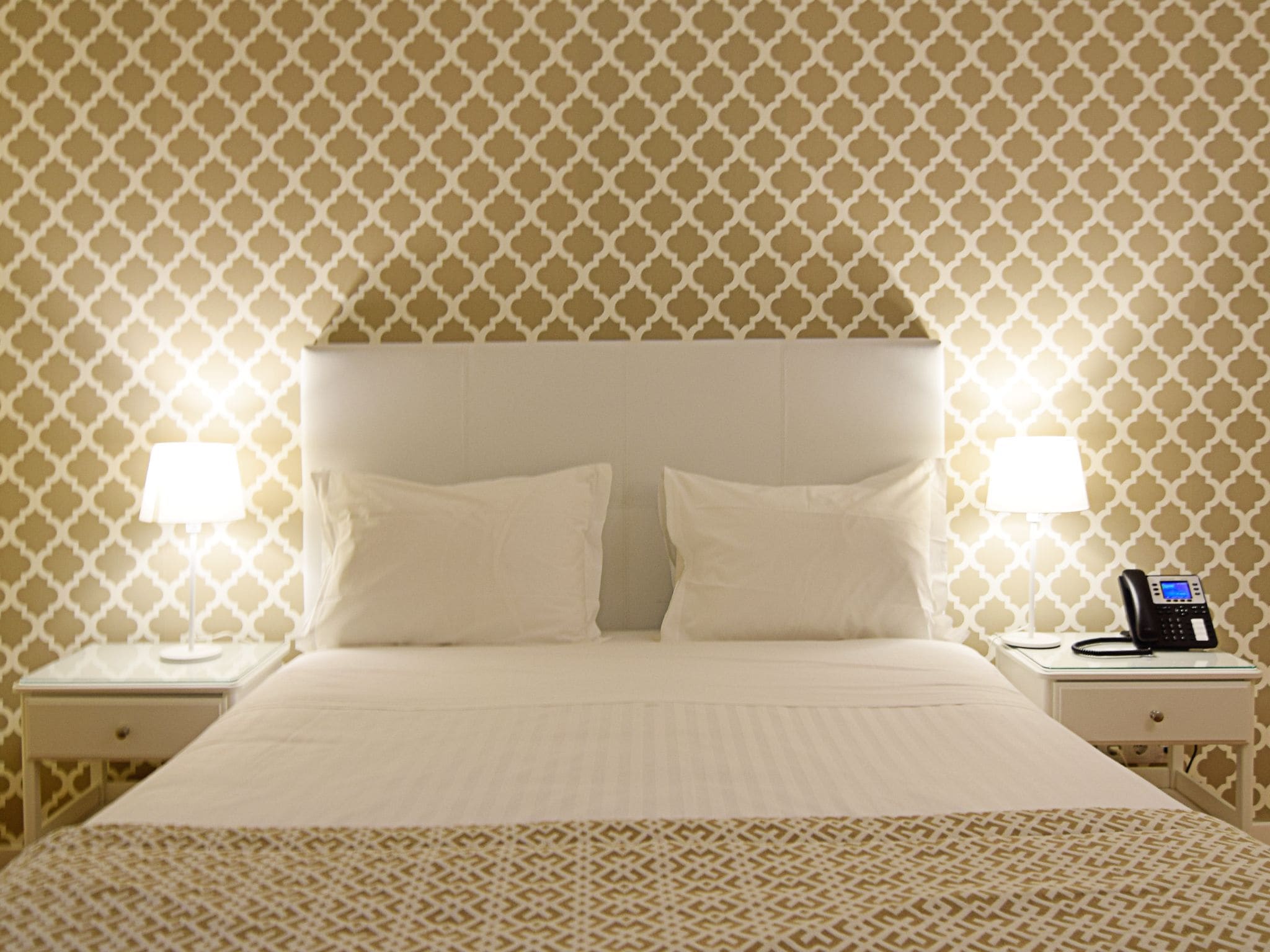 A hotel room with a patterned wallpaper, a bed with two pillows, two bedside lamps, and a telephone on the nightstand.