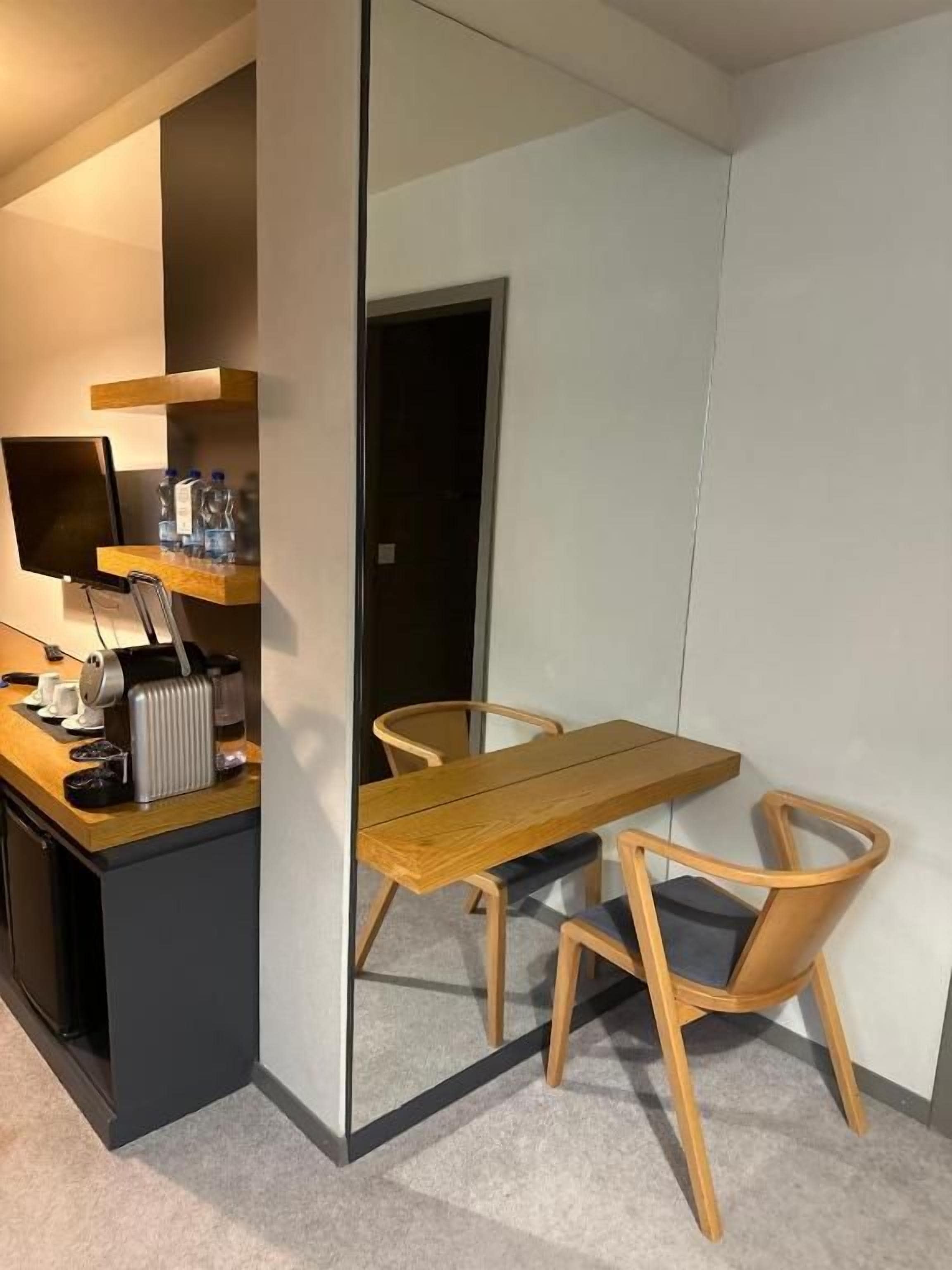 A compact room with a wooden dining table, two chairs, a small kitchen area with a toaster, and a mirror reflecting the room.