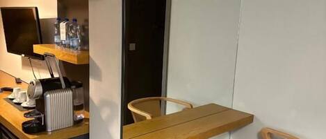 A compact room with a wooden dining table, two chairs, a small kitchen area with a toaster, and a mirror reflecting the room.