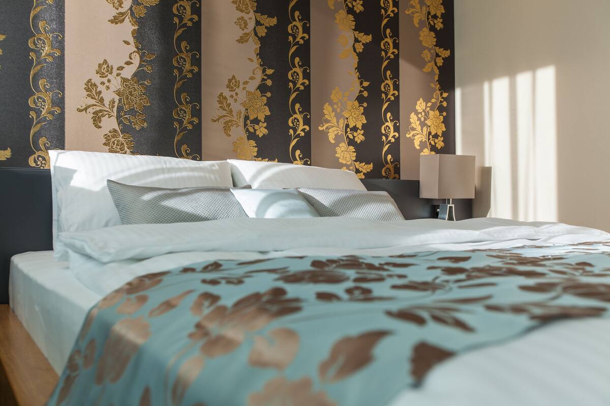 A neatly made bed with a floral-patterned bedspread, a bedside lamp, and a wall with vertical floral wallpaper.