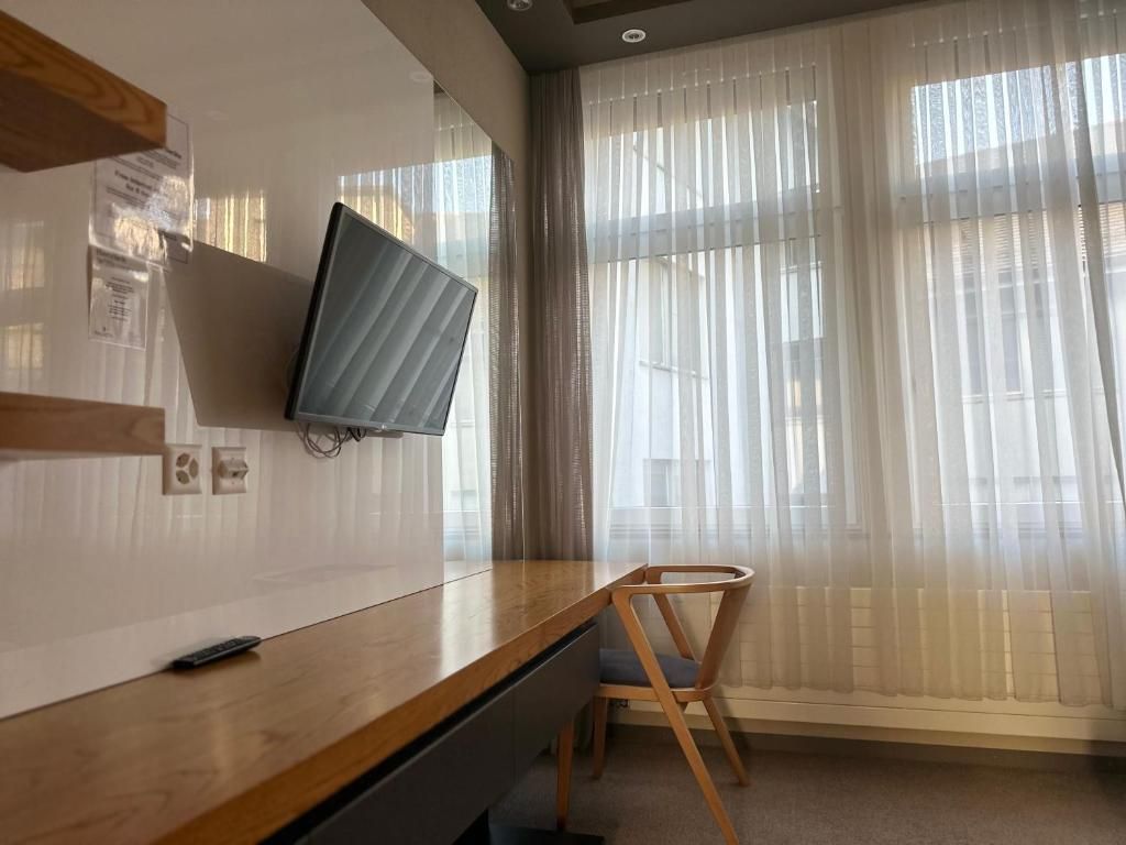 Une pièce avec un bureau en bois, une chaise, un téléviseur fixé au mur et une fenêtre ornée de rideaux légers.