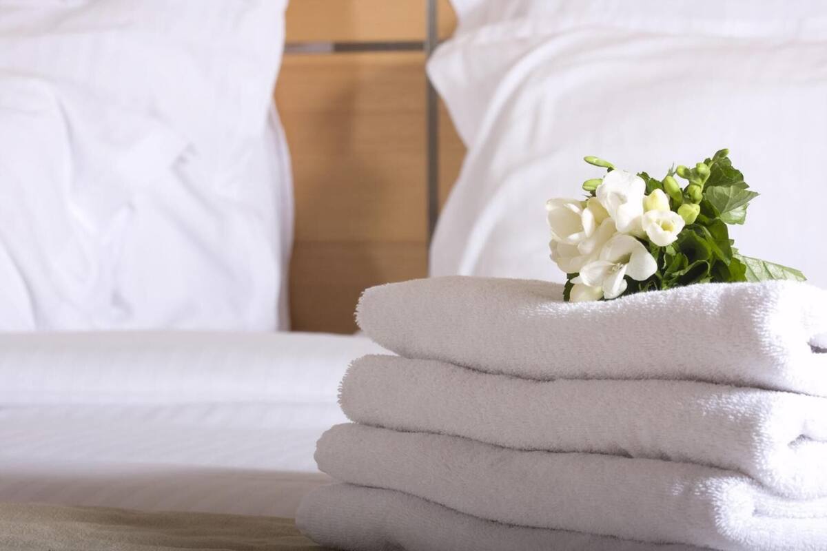 A stack of white towels on a bed with white bedding and a small bouquet of white flowers.