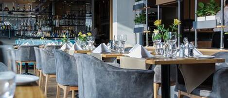 A restaurant interior with tables set for dining, grey upholstered chairs, and a bar area in the background.