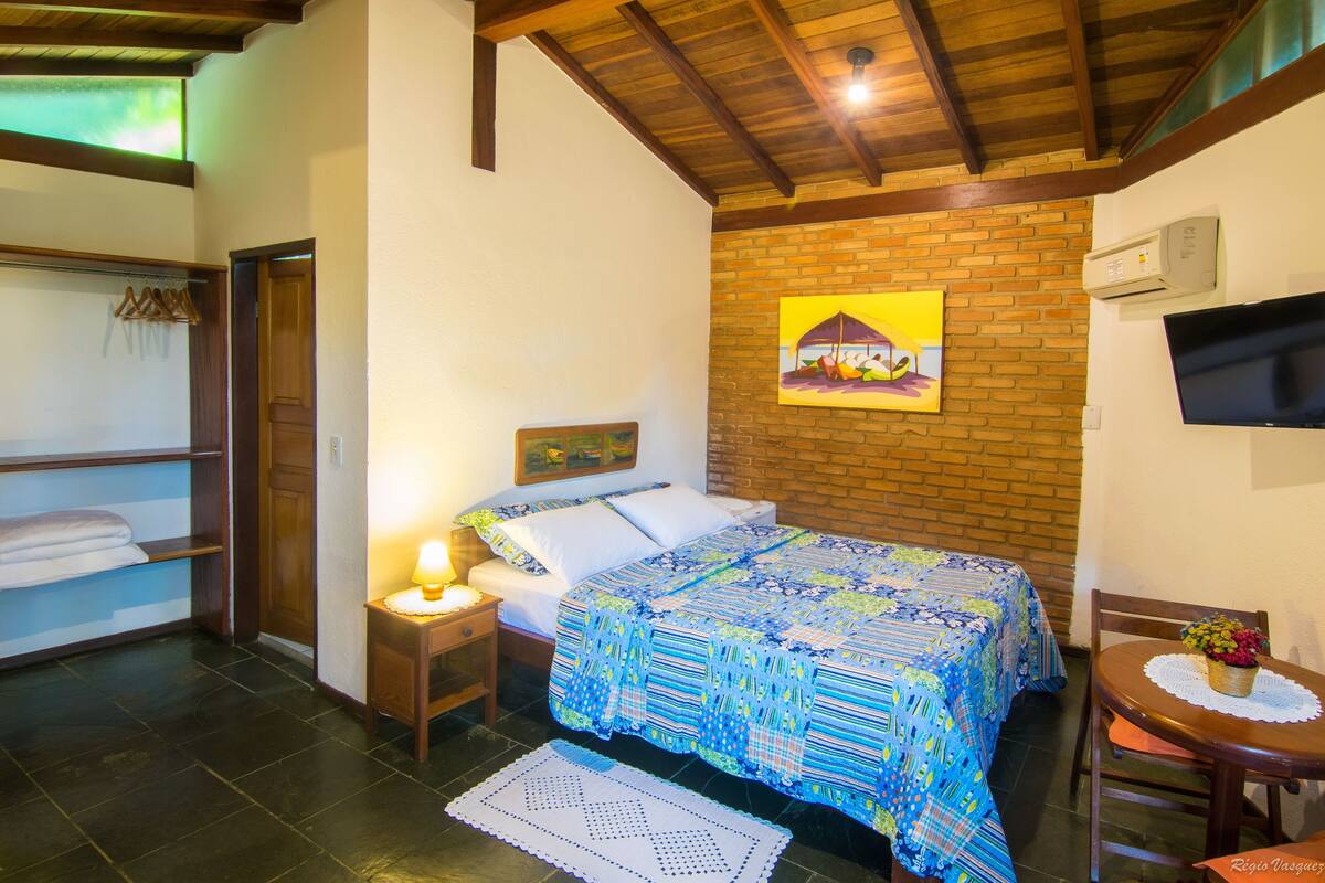 Un dormitorio con cama, mesita de noche con lámpara, una silla, una pequeña mesa con un florero y televisión en la pared.