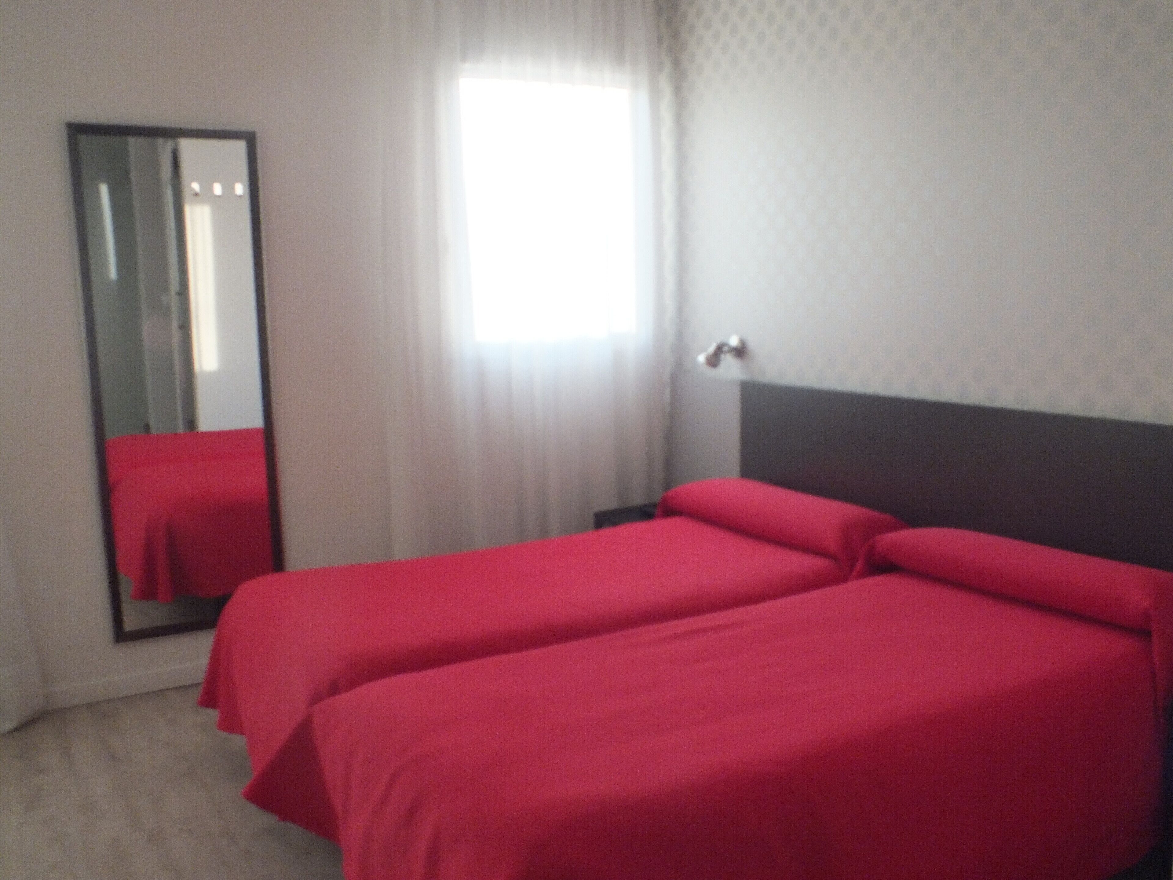 Habitación de hotel con dos camas individuales, un espejo grande, una ventana con cortinas traslúcidas y una pared lisa con textura.